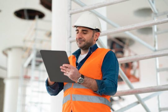 Construction supervisor wearing safety vest and helmet checking site details on a tablet beside scaffolding