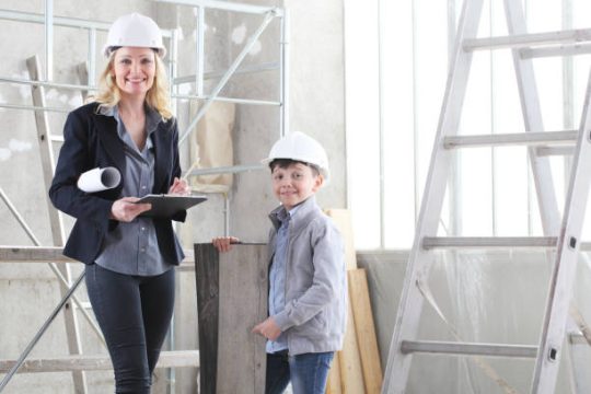 Female contractor and child wearing hard hats standing in an indoor renovation area with ladder and scaffold equipment