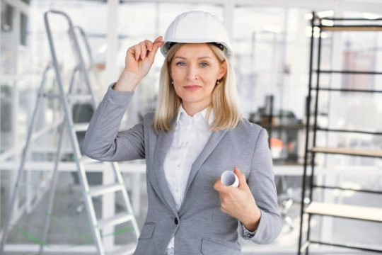Female construction professional wearing a hard hat and holding building plans inside a renovation site with scaffold behind her